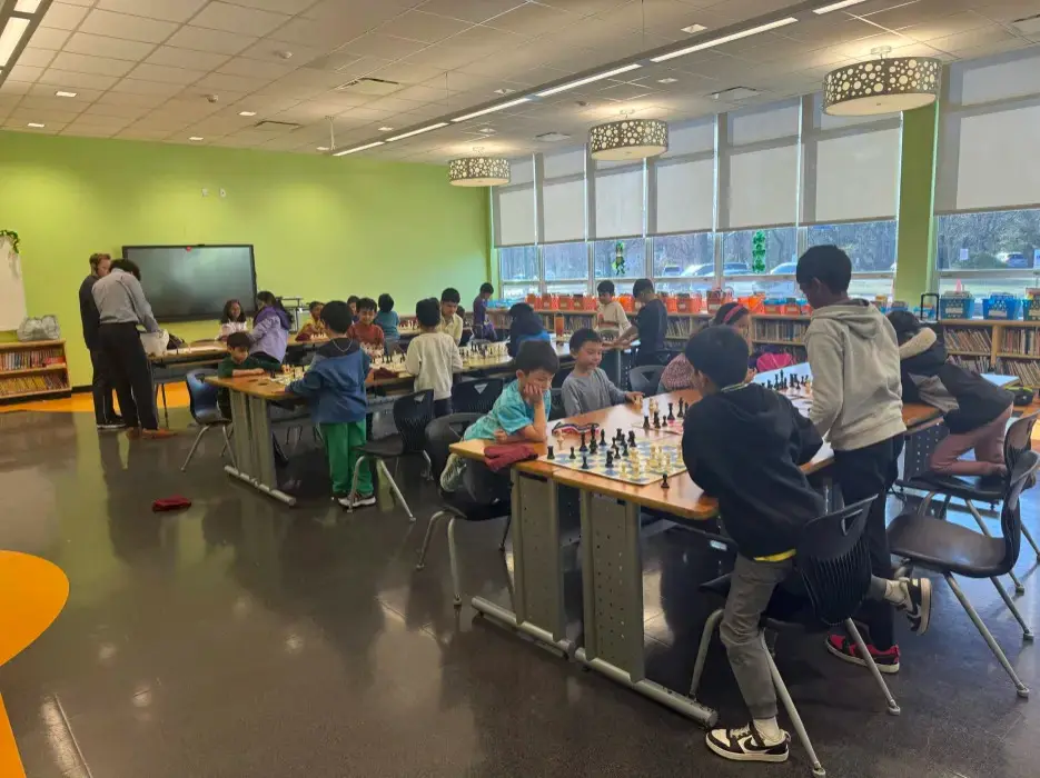 A wide view of a school library, students at long tables of chessboards under drum pendant lights.