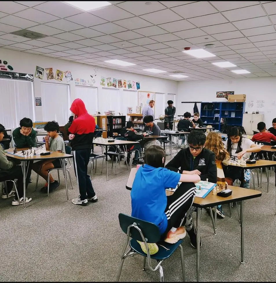 A wide classroom of students playing chess at desks under fluorescent ceiling panels.