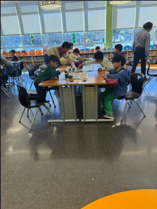 Students playing chess at a long library table, bookshelves behind them.
