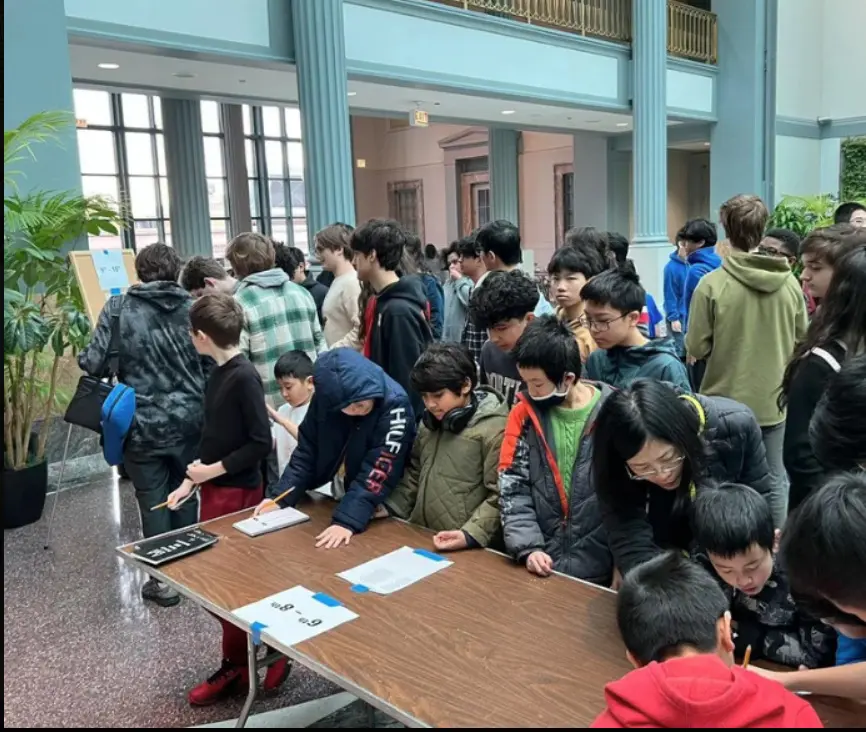 A crowd of students gathered around a long table, several writing on sheets of paper.