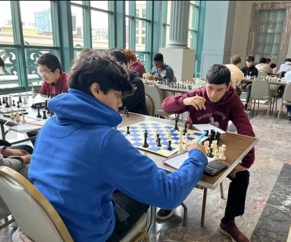Two students mid-game in a marble-columned hall, one considering his move with pen in hand.